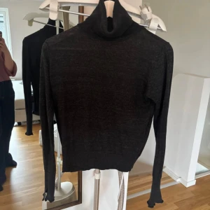 Svart polotröja i ull med slits vid ärm - Mörk, tunnstickad svart polotröja med lång ärm och cool slitsdetalj vid ärmslutet. Tröjan har en tight passform och hög polokrage, perfekt för lager på lager under höst och vinter.