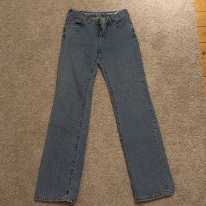 Blåa lågmidjade straight jeans 24/32 - Säljer ett par klassiska blåa jeans med låg midja och raka ben. Jeansen har fem fickor, bälteshällor och en snygg läderpatch bak i midjan. Perfekt passform för dig som gillar en avslappnad och tidlös look.