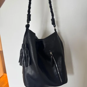 Svart axelväska med flätat axelband - Svart snygg axelväska med en snygg frans detalj. Defekt på baksidan - lite slitage inget som syns när den bärs då det är på baksidan.