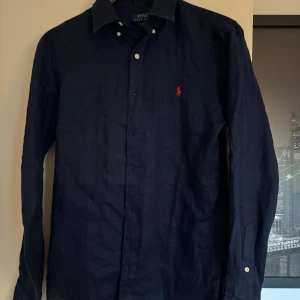 Mörkblå linneskjorta från Polo Ralph Lauren - Snygg mörkblå skjorta från Polo Ralph Lauren i 100% linne. Klassisk krage, knäppning framtill och ikonisk röd logga broderad på bröstet. Perfekt för en stilren och avslappnad look. Långärmad modell med vita knappar.