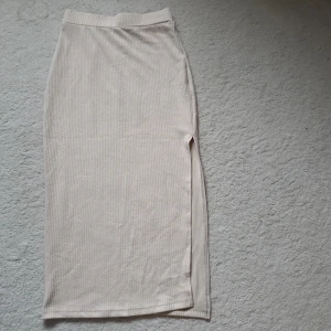 Beige ribbad långkjol med slits - Snygg beige långkjol i ribbat material med en stilren slits på sidan. Kjolen har en rak passform och bred resår i midjan för extra komfort. Perfekt för dig som gillar en enkel men trendig look.