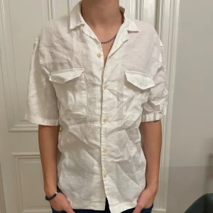 Vit linneskjorta från Zara - Storleken är S men passar M-L. Såhär sitter den på mig som är 180cm. Vit skjorta från Zara i luftig linne med kort ärm och två bröstfickor med lock. Klassisk krage och knäppning framtill. Perfekt för en chill och stilren look under varmare dagar.