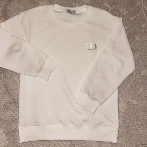 Vit sweatshirt från Christian Dior - Vit sweatshirt från Christian Dior med broderad CD-logga på bröstet. Tröjan har rund halsringning, ribbade muddar och är tillverkad i mjuk bomull. Perfekt för en clean och stilren look.