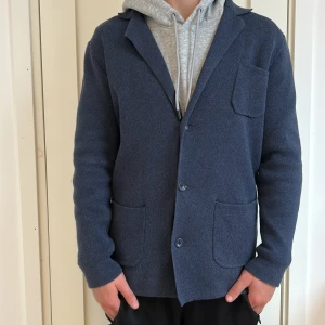 John Henric mörkblå stickad kavaj - Snygg mörkblå stickad kavaj med tre framfickor och knappar framtill. Klassisk krage och lång ärm, perfekt för en chill men ändå uppklädd look. Mjuk och bekväm känsla tack vare det stickade materialet.