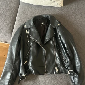 Svart skinnjacka från Zara - Säljer en svart skinnjacka från Zara med coola detaljer som dragkedjor, bälte i midjan och silvriga knappar. Jackan har klassisk bikerstil med bred krage och flera fickor framtill. Perfekt för dig som vill ha en edgy look.