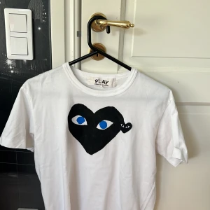 Vit Comme des Garçons Play t-shirt - Vit t-shirt från Comme des Garçons Play med det ikoniska svarta hjärtat med blå ögon tryckt på bröstet. Klassisk rund hals och korta ärmar. Perfekt för dig som gillar streetstyle och vill sticka ut med en cool detalj.