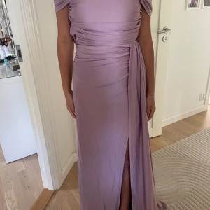 Ljuslila långklänning med slits - Säljer en elegant ljuslila långklänning med offshoulder-design och draperade detaljer runt midjan. Klänningen har en snygg slits framtill och ett mjukt, följsamt tyg som ger en lyxig känsla. Perfekt för dig som vill sticka ut med en feminin och modern look.