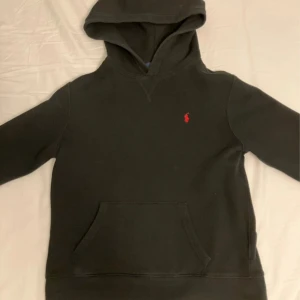 Svart hoodie från Polo Ralph Lauren - Svart hoodie från Polo Ralph Lauren med klassisk röd broderad logga på bröstet. Tröjan har huva, känguruficka och är tillverkad i mjuk bomull. Perfekt för dig som gillar stilrena och bekväma plagg.