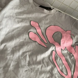 Grå t-shirt från syna world med rosa tryck - Säljer en grå t-shirt från syna world med stort rosa graffiti-inspirerat tryck på framsidan. T-shirten har rund hals och korta ärmar. Materialet är mjuk bomull och passformen är normal. Perfekt för dig som gillar streetwear och unika prints.