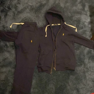 Svart hoodie från Polo Ralph Lauren S - Svart hoodie från Polo Ralph Lauren i storlek S. Klassisk modell med huva, dragkedja och vita snören. Broderad gul logga på bröstet och mjukt bomullsmaterial. Perfekt för chill dagar och enkel att matcha med allt.