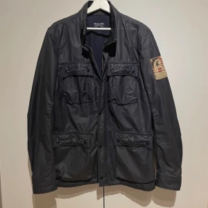 Ralph lauren Field jacka  - Svart jacka från Polo Jeans Company med fyra stora fickor framtill och patch med amerikansk flagga på ena ärmen. Jackan har dragkedja och knappar, klassisk krage och är tillverkad i slitstark bomull. Perfekt för dig som gillar en stilren och tidlös look. Storlek M men passar dig som har L
