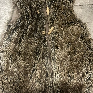 Brun pälsväst från Zara Girls - Säljer en fluffig brun och svart pälsväst från Zara Girls med snygga träknappar framtill. Västen har ett coolt mönster i olika bruna och svarta nyanser och är fodrad på insidan för extra komfort. Perfekt för att lyfta vilken outfit som helst.