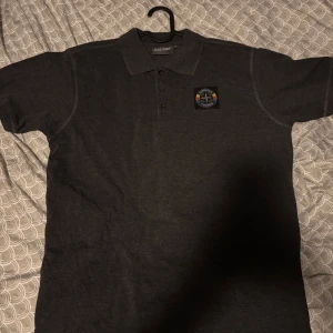 Djurgårds pike från open air 2018 - Svart pikétröja  i storlek S med Djurgården Stockholm-märke på bröstet. Klassisk krage, knäppning med tre knappar och korta ärmar. Perfekt för dig som vill visa ditt lagstöd med stil.