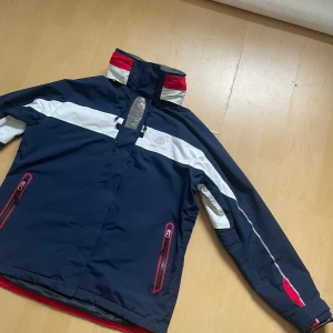  vindjacka från Henri Lloyd - Snygg marinblå vindjacka från Henri Lloyd med vita och röda detaljer. Jackan har hög krage, dragkedjor med röda kanter, och broderad logga på bröstet. 