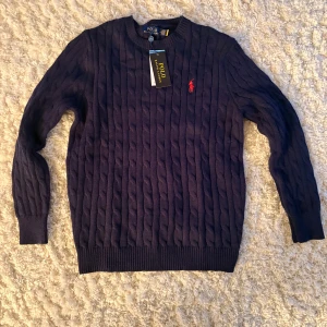 Mörkblå kabelstickad tröja Polo Ralph Lauren oanvänd  - Kabelstickad mörkblå tröja från Polo Ralph Lauren med klassisk rund halsringning och röd broderad logga på bröstet. Tröjan har långa ärmar och ribbade muddar för en snygg passform. Perfekt för dig som gillar stilren och tidlös design. Tröjan är i slim fit och har storleken M men skulle säga att den passar S mer än M