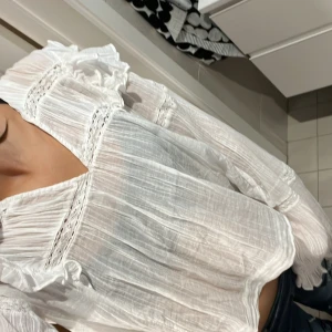 Vit blus med volang från Gina Tricot - Supersöt vit blus från Gina Tricot med volangdetaljer på axlarna och spetsinslag. Blusen har v-ringning, långa ärmar och är tillverkad i ett tunt, luftigt bomullstyg. Perfekt för dig som gillar romantisk och bohemisk stil. 