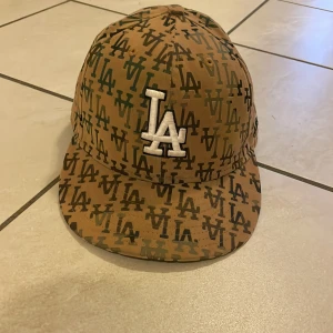 LA keps från New Era 59FIFTY, beige - Säljer en beige New Era 59FIFTY keps med LA-logga i vitt framtill och all-over print med svarta och gröna LA-bokstäver. Klassisk flat brim och broderad MLB-logga bak. Perfekt för dig som gillar streetstyle och sportig look.