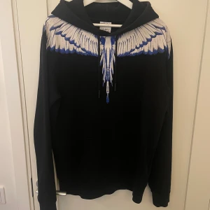Marcelo Burlon svart hoodie med vingar - Svart hoodie från Marcelo Burlon med ikoniskt vingtryck i vitt och blått över axlar och bröst. Tröjan har huva med snörning och en klassisk känguruficka framtill. Perfekt statement-plagg för dig som vill sticka ut.