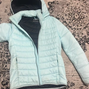 Ljusblå pufferjacka från Everest - Säljer en ljusblå pufferjacka från Everest med huva och dragkedja. Jackan är quiltad och har svarta detaljer på insidan. Perfekt för kyliga dagar och har en snygg, sportig look.