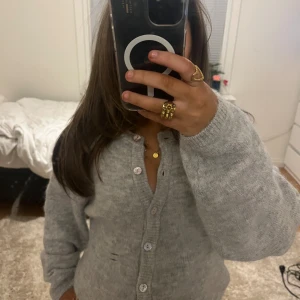 Grå stickad kofta med knappar - (Reporna är i spegeln) Mysig grå stickad kofta med pärlemorknappar och ribbade muddar. Koftan har en klassisk passform och är perfekt att slänga på sig när det är kyligt. Enkel och stilren design som funkar till allt.🩶