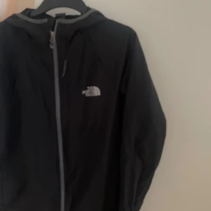 Svart vindjacka från The North Face - Svart vindjacka från The North Face med huva och hel dragkedja framtill. Jackan har en enkel och sportig design med logga på bröstet. Perfekt för blåsiga dagar och outdoor-aktiviteter. 