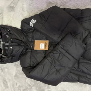 Svart pufferjacka från Burberry - Svart pufferjacka från Burberry med klassisk logga på axeln och rutigt innerfoder i huvan. Jackan har dragkedja framtill och är vadderad för extra värme. Tillverkad i bomull och polyamid, perfekt för kalla dagar. Snygg och tidlös design med streetkänsla.