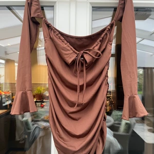 Brun omlottklänning med volangärm - Supersnygg brun kortklänning med omlottdesign och långa ärmar med volang vid ärmslut. Klänningen har snörning framtill och en draperad effekt som ger en cool och trendig look. Perfekt för dig som gillar unika detaljer och vill sticka ut.