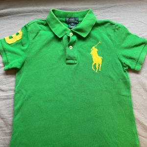 Grön pikétröja från Ralph Lauren - Snygg grön pikétröja från Ralph Lauren med klassisk krage, tre knappar och stor gul broderad logga på bröstet. Pikén har korta ärmar och en gul detalj på ena ärmen. Perfekt för en sportig och fräsch look.