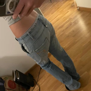 Ljusblå lågmidjade jeans med broderad ficka - Säljer ett par ljusblå jeans från Denny Rose med snygga slitningar och broderad detalj på bakfickan. Modellen har låg midja och lite utsvängda ben med en relaxed fit. Klassiska femficksjeans i mjukt denimtyg, perfekta för en chill och trendig look. innerbenslängd 84cm, midjemått 38cm