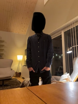 Mörkblå skjorta från Ralph Lauren - Klassisk mörkblå skjorta från Ralph Lauren med button-down krage och broderad logga på bröstet. Skjortan har långa ärmar och vita knappar framtill. Perfekt till jeans eller chinos för en clean och stilren look.