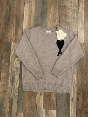 Grå stickad tröja från Ami - Ljusgrå stickad tröja från Ami med rund halsringning och ribbade muddar. På bröstet finns ett svart hjärta ovanför ett stort A, vilket ger en unik och trendig look. Perfekt för dig som gillar stilrena och moderna plagg.