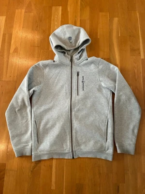 Ljusgrå hoodie från Sail Racing - Snygg ljusgrå hoodie från Sail Racing med dragkedja framtill och huva. Hoodien har två sidofickor och en bröstficka med dragkedja. Tillverkad i mjukt bomullsmaterial och har diskret logga på bröstet. Perfekt för en avslappnad och sportig stil.