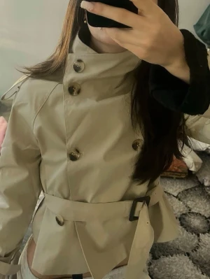 Beige trenchcoat med bälte - Stilren beige trenchcoat med klassisk dubbelknäppning och bälte i midjan. Jackan har stora knappar, axelklaffar och hög krage. Perfekt för dig som vill ha en tidlös och trendig look.