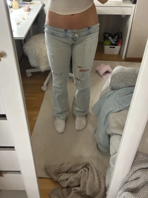 Ljusblå bootcut jeans med slitningar - Säljer ett par väldigt lågmidjade ljusblå bootcut jeans med slitna detaljer och hål på benen. Jeansen har låg midja och klassisk femficksdesign. Perfekta för dig som gillar en avslappnad och trendig look. Personen i bilden är 171