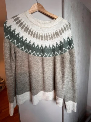 Beige stickad tröja från Vero Moda - Stickad tröja från Vero Moda i beige och vitt med grönt och brunt mönster runt oket. Tröjan har rund hals, långa ärmar och ribbade muddar vid ärmslut och nederkant. Perfekt för kalla dagar och riktigt mysig att bära.