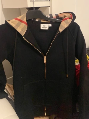 Svart Burberry hoodie med dragkedja - Snygg svart hoodie från Burberry med klassiskt rutigt foder i huvan. Dragkedja framtill, fickor och guldfärgade detaljer på dragkedjan. Perfekt för dig som vill ha en stilren men ändå lyxig hoodie med ikoniskt mönster i huvan.