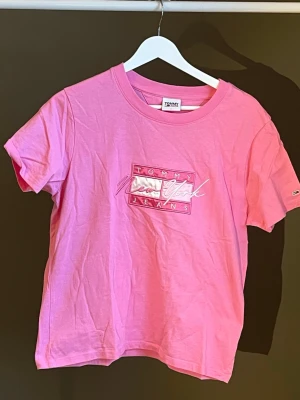 Rosa t-shirt från Tommy Jeans - Snygg rosa t-shirt från Tommy Jeans med tryck på bröstet där det står 'Tommy New York Jeans'. Klassisk rund hals och korta ärmar. Tillverkad i mjuk bomull som känns skön mot huden. Perfekt för en avslappnad och trendig look. Köpt from Tommy Hilfiger, aldrig använt🌸