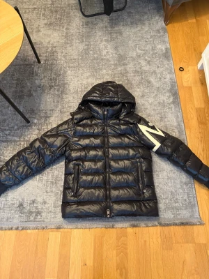 Svart dunjacka från Moncler - Svart puffer dunjacka från Moncler med huva och dragkedja framtill. Jackan har två fickor med dragkedja och en stor vit Moncler-logga på ena ärmen. Perfekt för kalla dagar och riktigt snygg streetstil.
