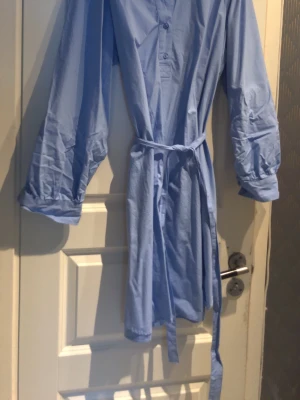 Blå skjortklänning från B. Copenhagen - Snygg blå skjortklänning från B. Copenhagen med knappar framtill och matchande knytskärp i midjan. Klänningen har lång ärm och en avslappnad passform, perfekt för en trendig och enkel look.