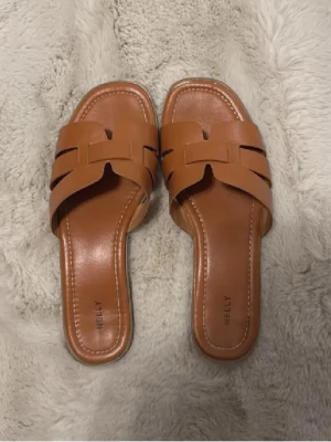 Bruna sandaler från Nelly - Snygga bruna sandaler från Nelly med öppen tå och platt sula. De har ett flätat band över foten i syntetmaterial och en stilren design som passar perfekt till sommaren.