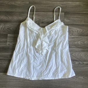 Vit linnetopp med volang från Atmosphere - Söt vit linnetopp från Atmosphere gjord i 100% bomull. Storlek 40 men passar mig som bär s bra.Toppen har smala axelband som är justerbara, v-ringad hals och snygg volangdetalj framtill. Perfekt för varma dagar när man vill vara lite extra fin. Rekommenderar att ha vita kläder under då man kan se igenom i vissa ljus. Har tyvärr ingen koll på orginalpriset. (Pris kan diskuteras)