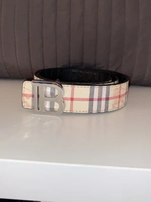 Burberry rutigt bälte med B-spänne - Snyggt bälte från Burberry med klassiskt beige, rött och svart rutmönster. Bältet har ett stort silvrigt B-spänne framtill och är gjort i läder med canvasdetaljer. Perfekt accessoar för att lyfta din outfit.