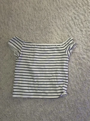 Randig offshoulder topp från H&M - Säljer en vit offshoulder topp från H&M med svarta horisontella ränder. Toppen har korta ärmar och är croppad i modellen. Perfekt för en avslappnad och trendig look. Materialet är mjukt och stretchigt, troligtvis bomull med lite syntet.