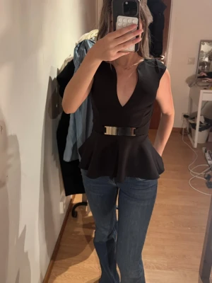 Svart peplumtop med guldbälte - Svart topp med djup V-ringning, peplum och metallbälte från Nelly. Har aldrig haft den då den är för liten. Helt ny, men saknar prislapp (fick den som present).  Några repor på metallbälte, inget som syns.