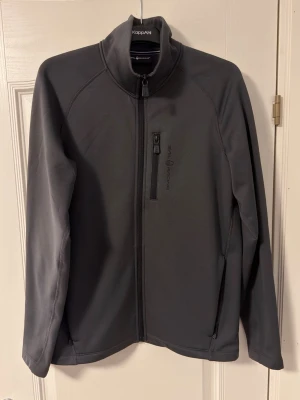 Tröja från Sail Racing - Mörkgrå ziptröja från Sail Racing med hög krage och dragkedja framtill. Tröjan har en bröstficka med dragkedja och diskret logga på bröstet. Tillverkad i ett stretchigt funktionsmaterial som passar perfekt till lager-på-lager.