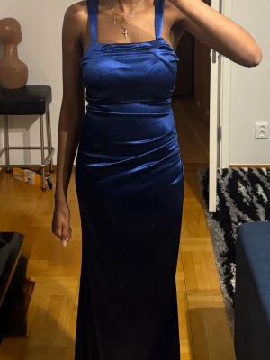 Marinblå långklänning i satin - Säljer en elegant marinblå långklänning i glansigt satinmaterial. Klänningen har breda axelband, figurnära passform och snörning i ryggen för en snygg siluett. Perfekt för dig som vill sticka ut med en stilren look.