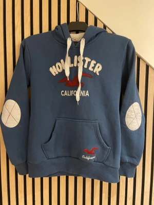 Blå Hollister hoodie med tryck - Blå hoodie från Hollister med vit broderad text och röd logga på bröstet. Har vita dragsnören i huvan, känguruficka och gråa armbågslappar med röda detaljer. Mjuk insida och ribbade muddar. Perfekt för en avslappnad stil. Storleken är märkt M (Medium) i tröjan men den är mer som en S/XS (Small/Xtra Small) i storleken. Tröjan är i ett väldigt bra skick. 