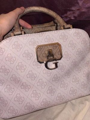 Beige handväska från Guess - Snygg beige handväska från Guess med logomönster och guldfärgade detaljer. Väskan har ett robust handtag och ett stort G-spänne framtill. Perfekt för dig som vill ha en trendig och praktisk accessoar med plats för det viktigaste.