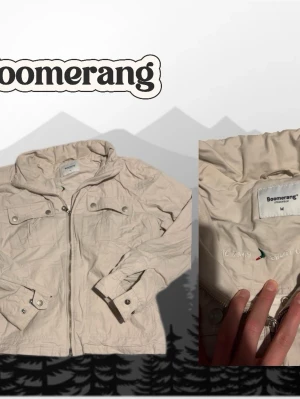 Beige vindjacka från Boomerang - Snygg beige vindjacka från Boomerang med dragkedja framtill, fyra fickor med knappstängning och huva. Tillverkad i lätt polyester, perfekt för blåsiga dagar. Jackan har en klassisk passform och stilrena detaljer.