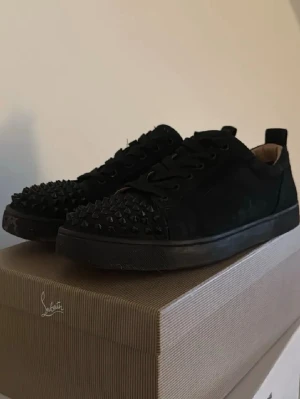 Loubs skor  - Säljer ett par svarta sneakers från Christian Louboutin med mattsvarta nitar på tån och klassisk röd sula. Skorna har snörning och är tillverkade i mocka för en lyxig känsla. Perfekta för dig som vill sticka ut med en edgy och exklusiv look. Kan även byta imot en klocka om du kan mötas i Jönköping 
